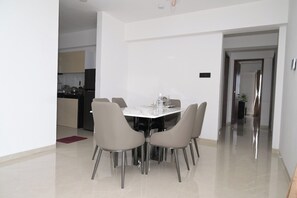 A modern dining area with a white table and four chairs, a kitchen in the background, and a hallway leading to another room. - Hotel Nestoria Corporate Services Aparts (Pune)
