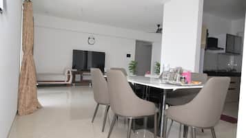A modern dining area with a round table, four chairs, a clock on the wall, and a kitchen in the background.