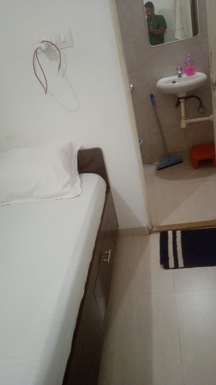 A room with a bed, a white wall socket, a mirror, a sink, and a mop.