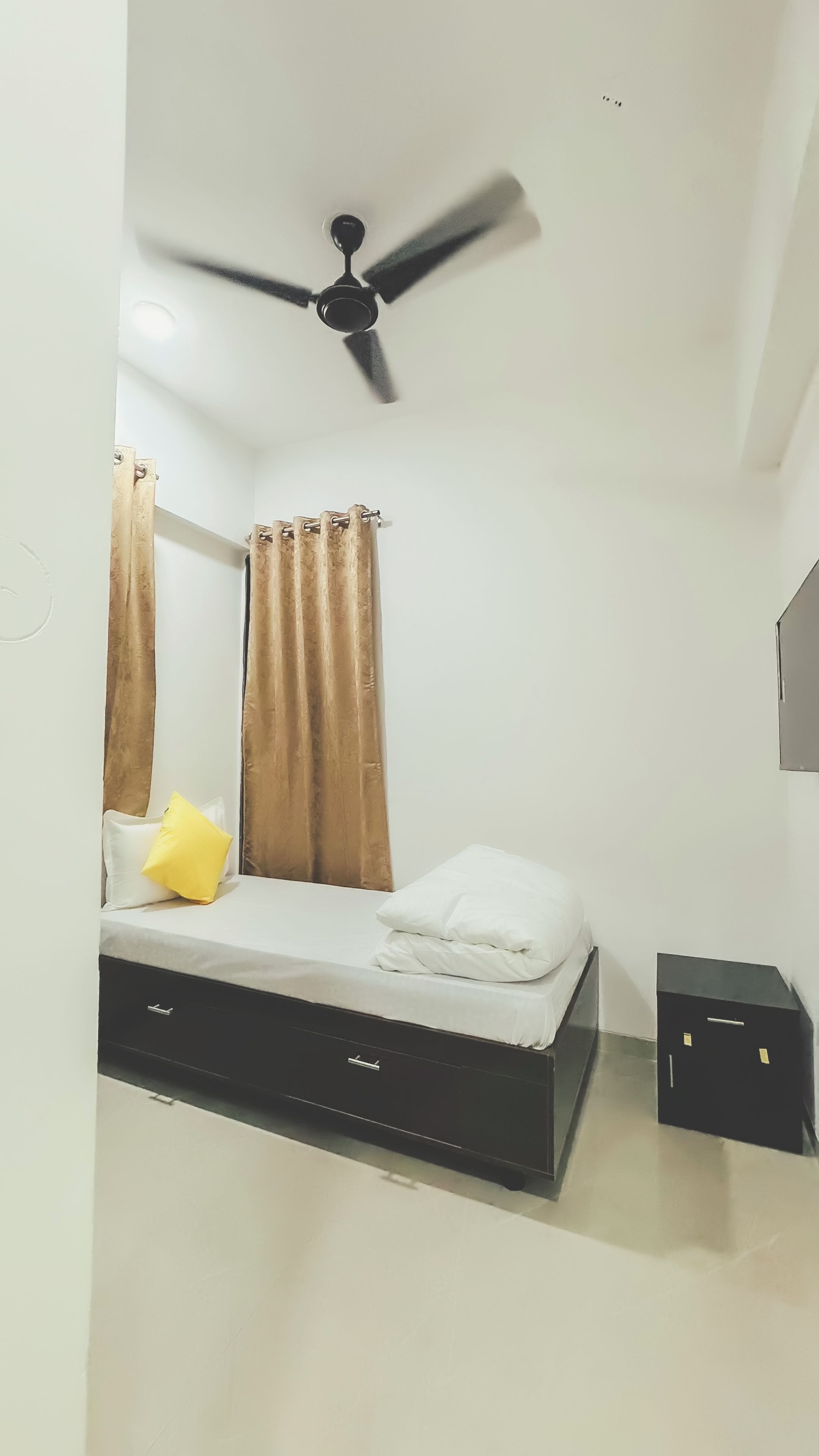 Une chambre à coucher avec un lit, des rideaux, un ventilateur de plafond et une petite armoire noire.
