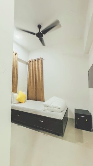 A bedroom with a bed, curtains, a ceiling fan, and a small black cabinet. - Hotel Nestoria Corporate Services Aparts (Pune)