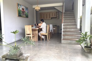 Un espace repas avec une personne assise à une table, des chaises en bois, une plante en pot et un escalier menant à un autre niveau.