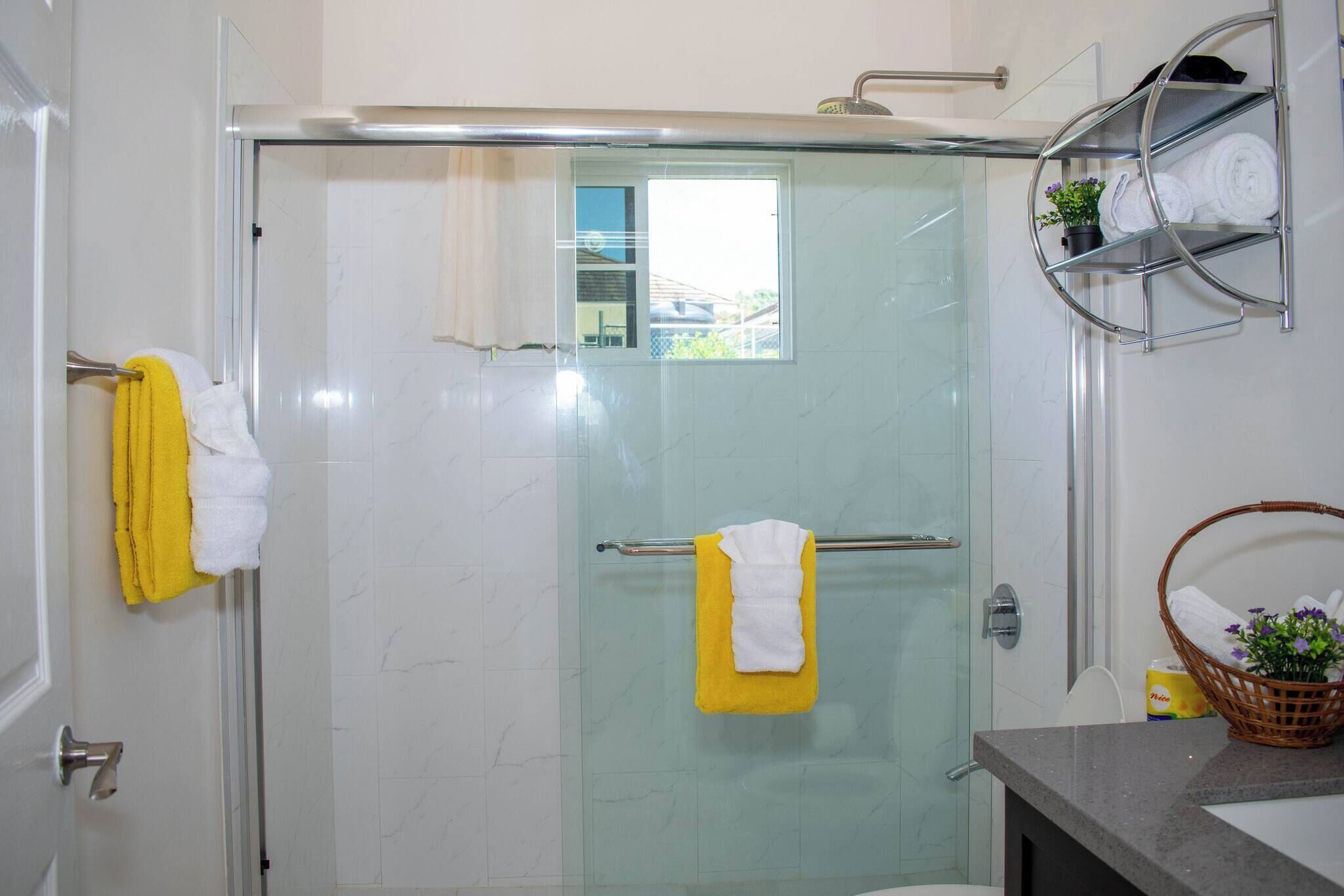 A bathroom with a glass shower enclosure, a towel rack with yellow and white towels, a sink with a basket of toiletries, and a small window.