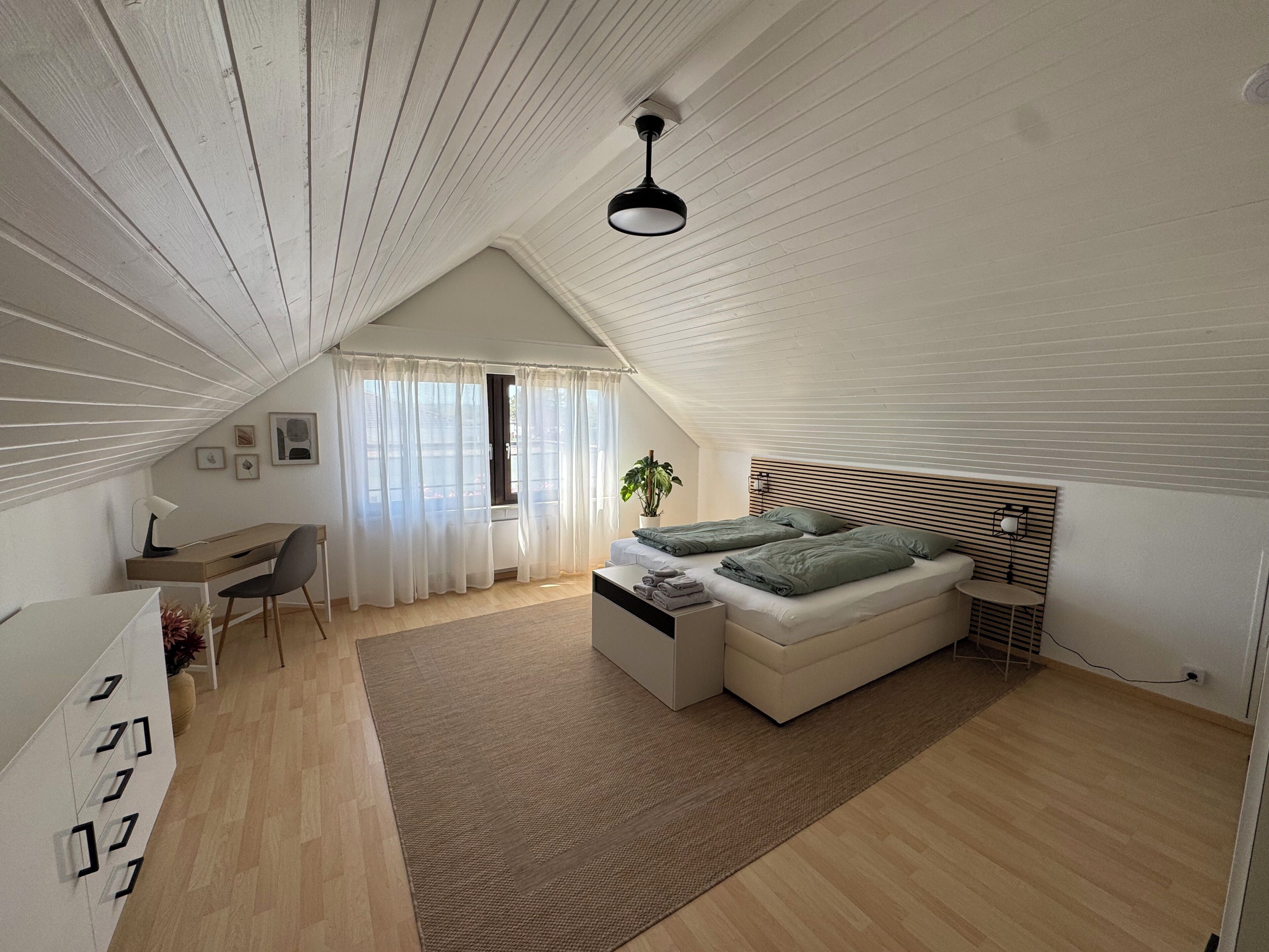 A modern bedroom with a sloped ceiling, a bed with green bedding, a desk with a chair, and a small table with a lamp.