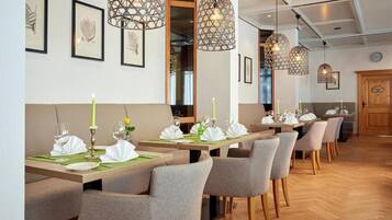 Ein Restaurant mit langen Tischen, grauen Stühlen und hängenden Pendelleuchten.