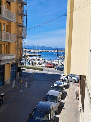 A street view with parked cars, a marina with boats, and a building with a balcony. - A View of the Harbor (Monte Argentario)
