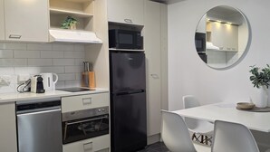 Eine kleine Küche mit einem schwarzen Kühlschrank, einer Mikrowelle, einer Kaffeemaschine und einem Spülbecken. Ein Esstisch mit Stühlen und ein Spiegel sind an der Wand angebracht.