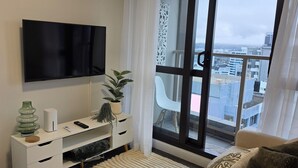 Ein modernes Wohnzimmer mit Fernseher, einem weißen Unterhaltungsschrank, einer Topfpflanze und Blick auf die Stadt durch große Fenster.