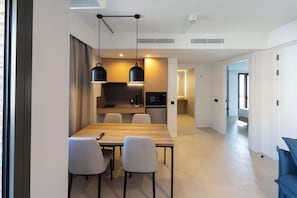 A modern dining area with a wooden table and chairs, overhead pendant lighting, and a kitchen in the background.