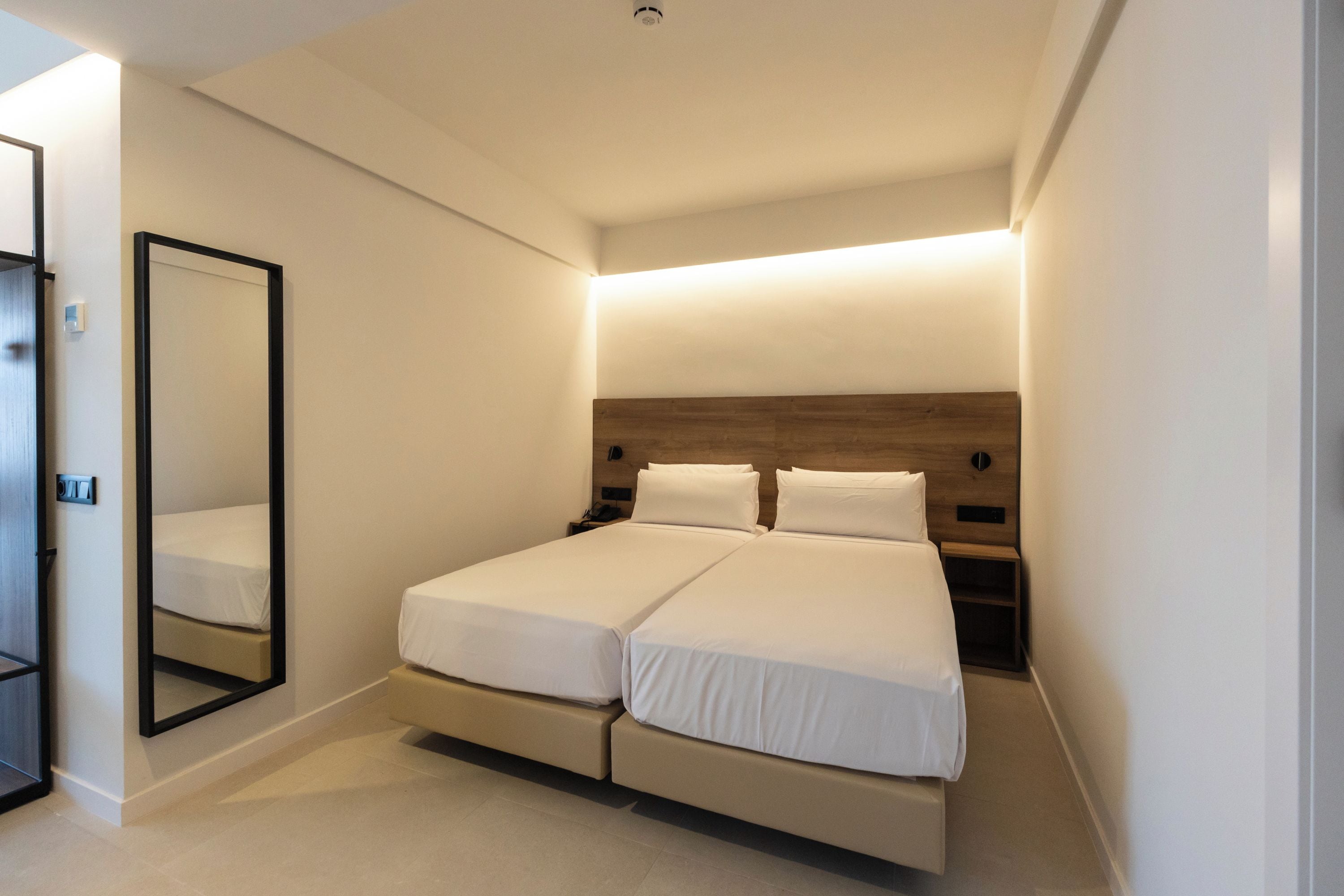 Quarto de hotel moderno com uma cama de casal, mesa de cabeceira, espelho e cabeceira de madeira.