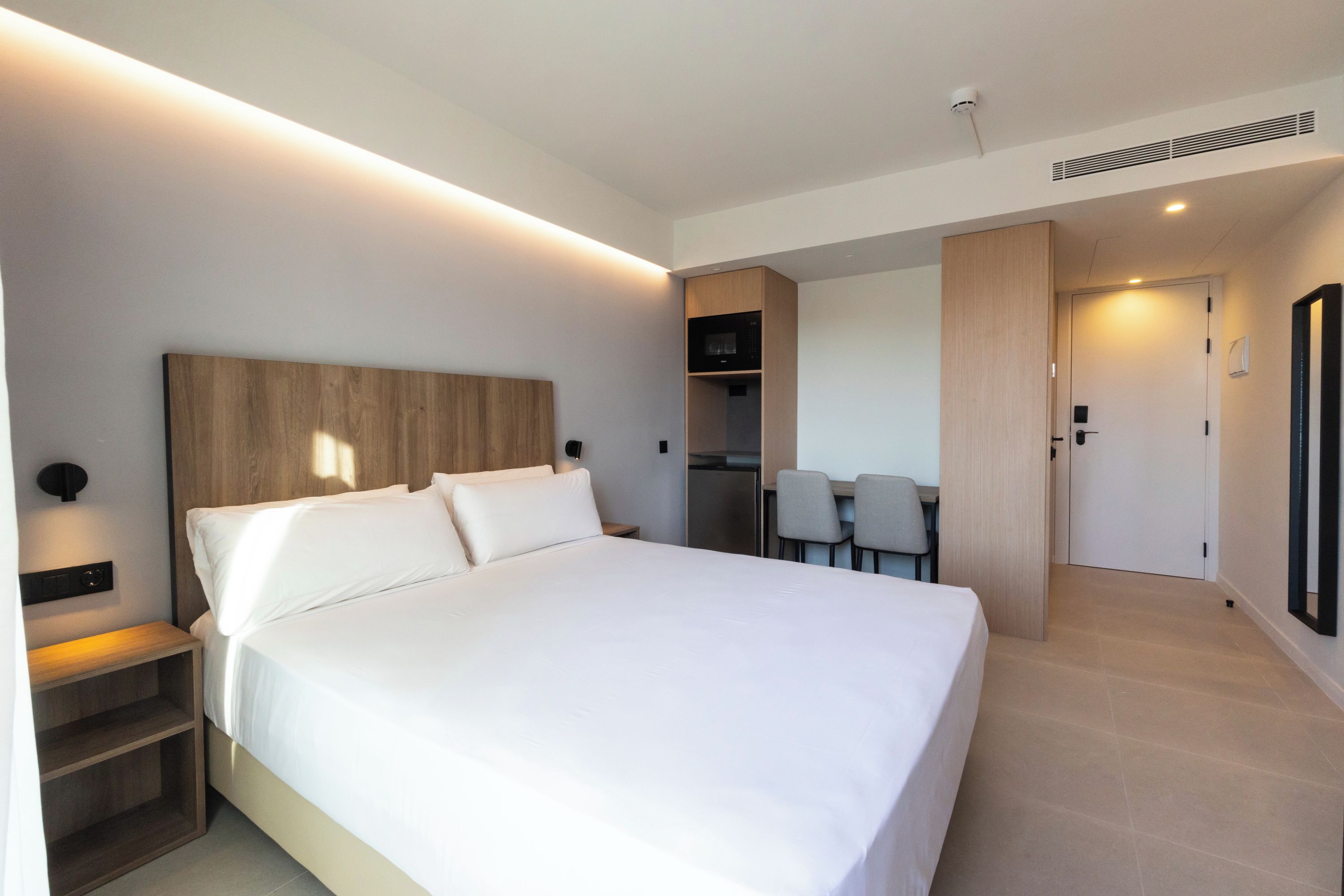 Habitación de hotel moderna con una cama grande, una mesita de noche, un armario empotrado y una pequeña cocina.