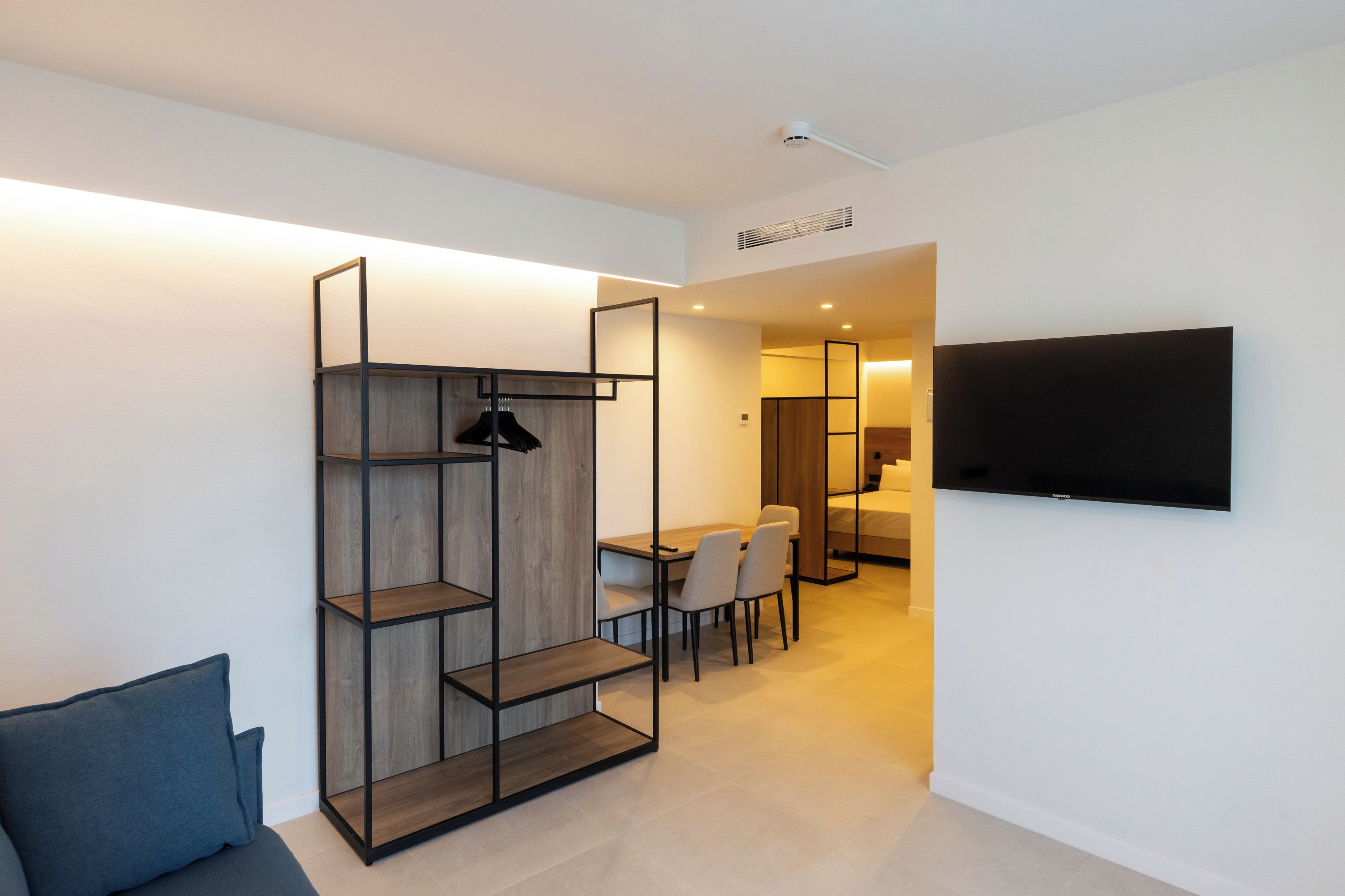 Ein modernes Hotelzimmer mit einer eingebauten Holzwandregal, einem Wand-Fernseher, einem Essbereich mit Tisch und Stühlen und einem separaten Schlafbereich mit Bett.