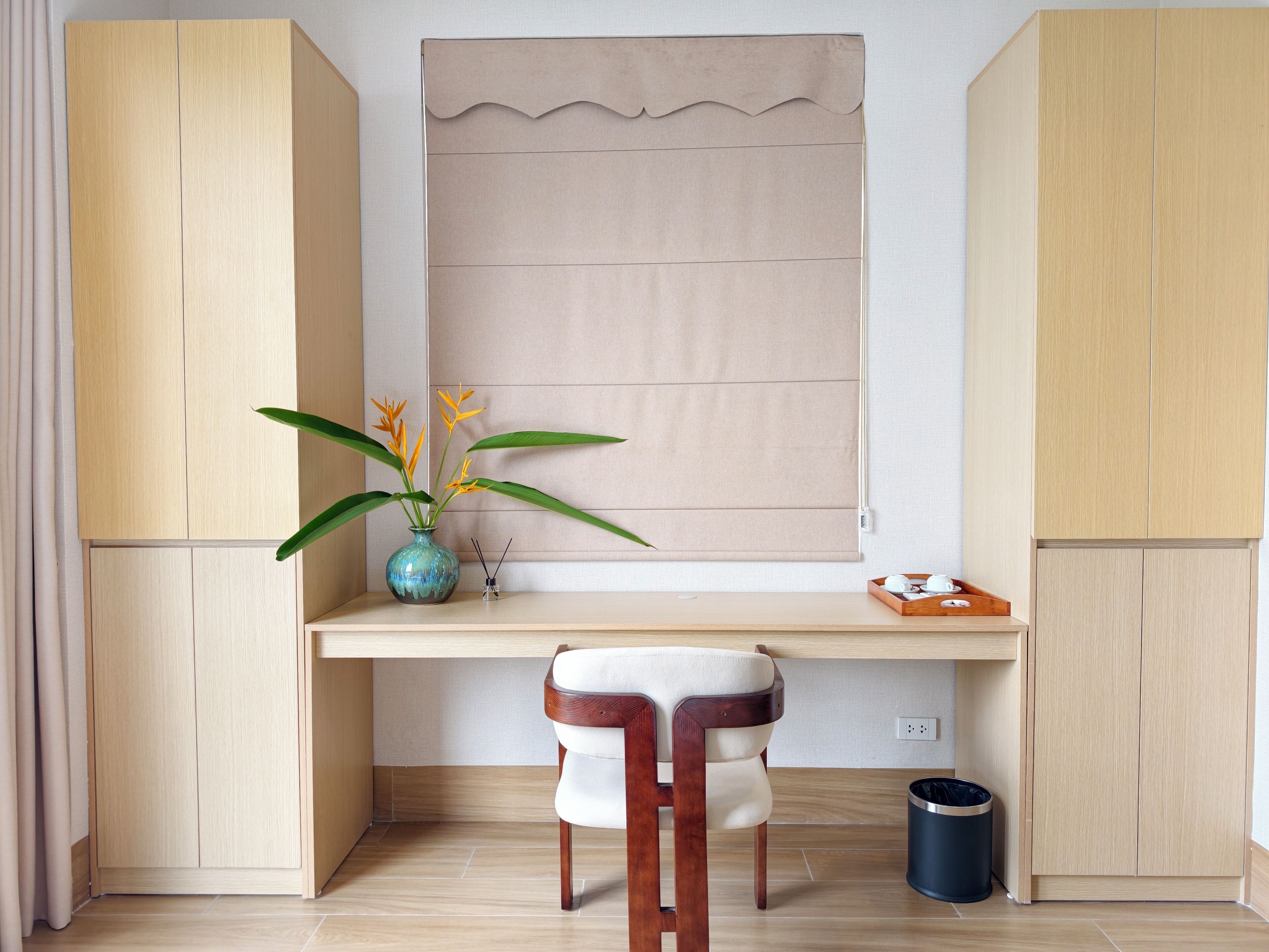 A room with a wooden desk, a chair, a vase with flowers, and a window with a blind.