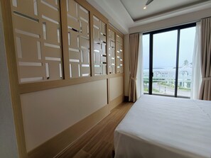 Een hotelkamer met een groot bed, houten vloer, uitzicht op zee en een raam met gordijnen.