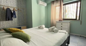 A neatly made bed with pillows, a bedside table with towels, a hanging rack with clothes, and a window with curtains. - Mondello Bay - Roosevelt Apartments (Palermo)