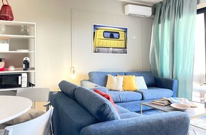 A living room with a blue sofa, a small table, a white chair, a bookshelf, and a wall-mounted picture. - Mondello Bay - Roosevelt Apartments (Palermo)