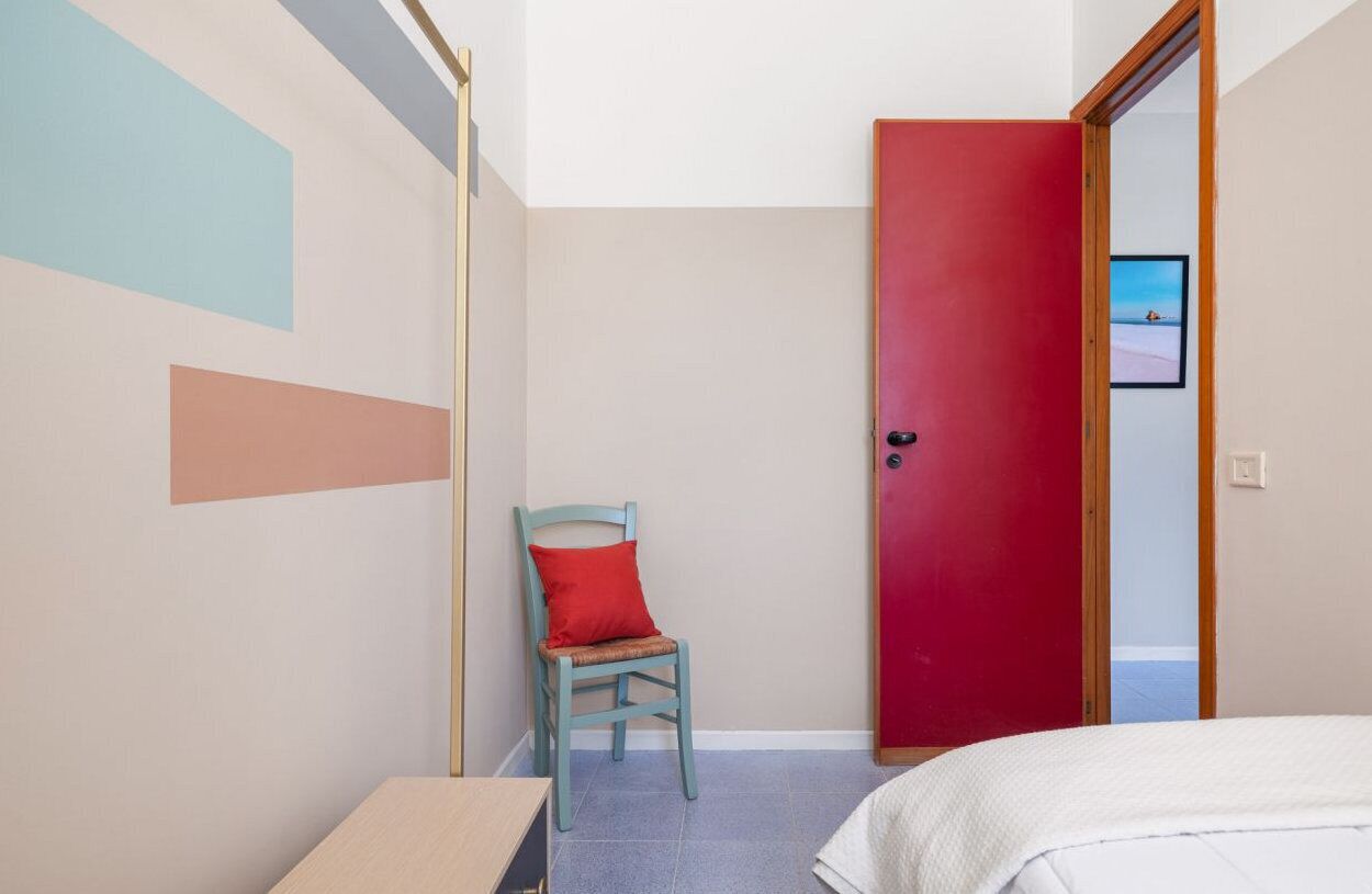 A bedroom with a bed, a chair with a red cushion, a red door, and a wall with geometric shapes.
