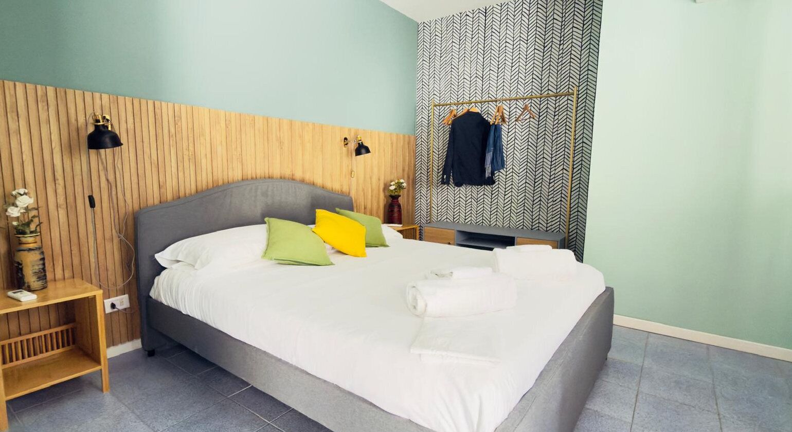 Una cama bien tendida con almohadas, una mesita de noche con una lámpara y un perchero frente a una pared con diseño.