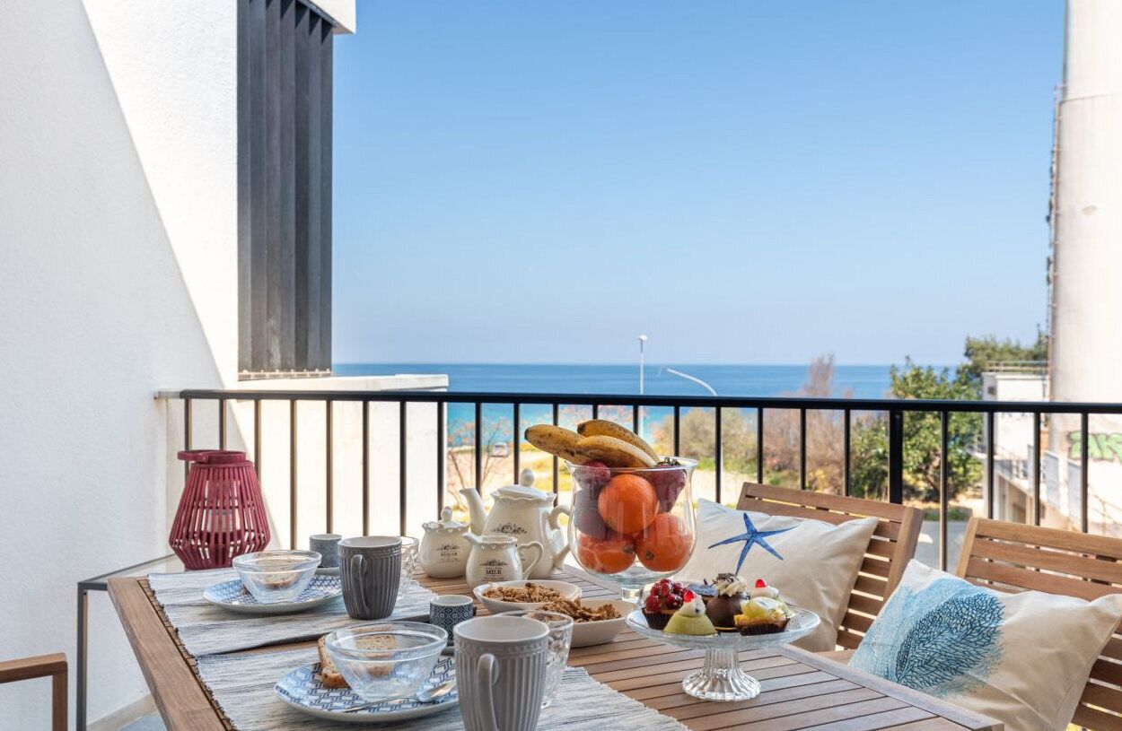 A balcony with a table set for breakfast, including a fruit bowl, pastries, and a teapot.
