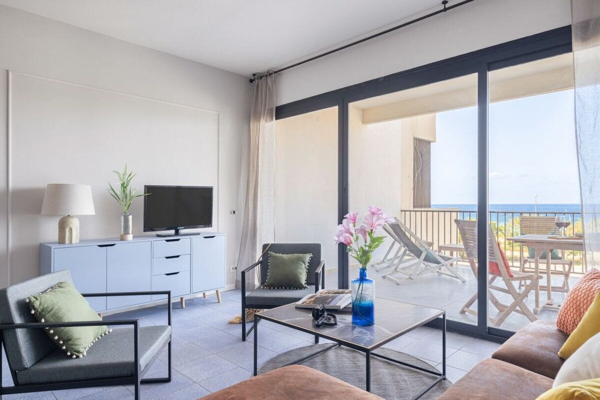 A modern living room with a sofa, armchair, coffee table, and a television on a cabinet. There is a balcony with outdoor seating and a view of the sea.