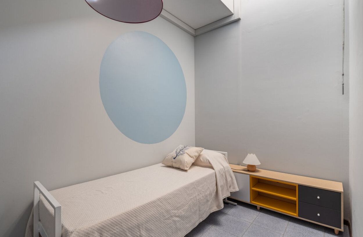 A small bedroom with a single bed, a bedside table with a lamp, a wall-mounted shelf, and a large circular wall decoration.