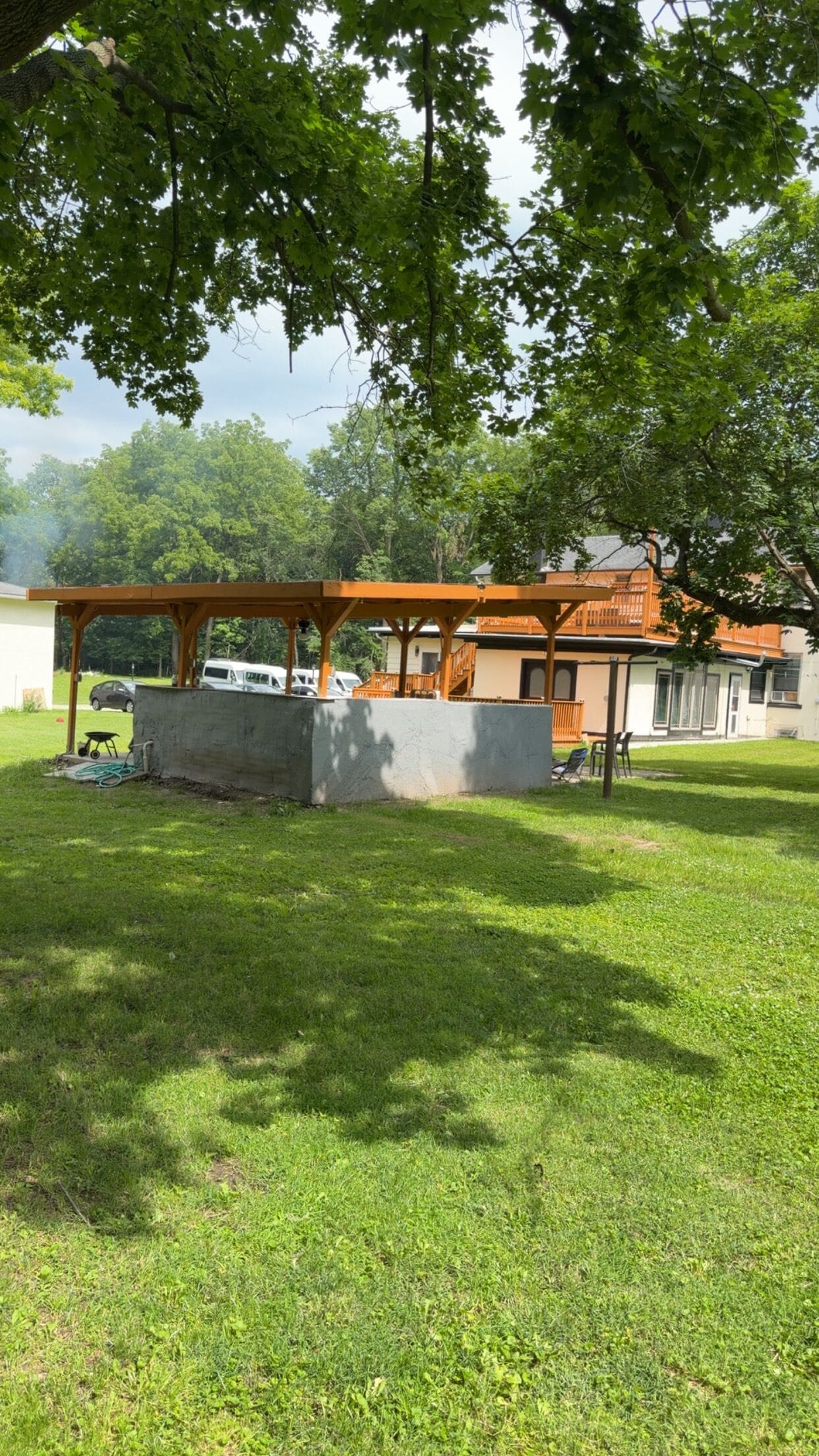 Un'area esterna coperta con struttura in cemento, una pergola in legno e un edificio sullo sfondo.