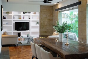 Un salon moderne avec une table à manger en bois, des canapés blancs, un téléviseur et une bibliothèque.