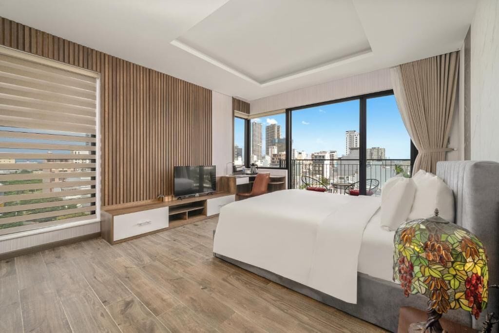 A modern hotel room with a large bed, a desk, a TV, and a balcony with a city view.