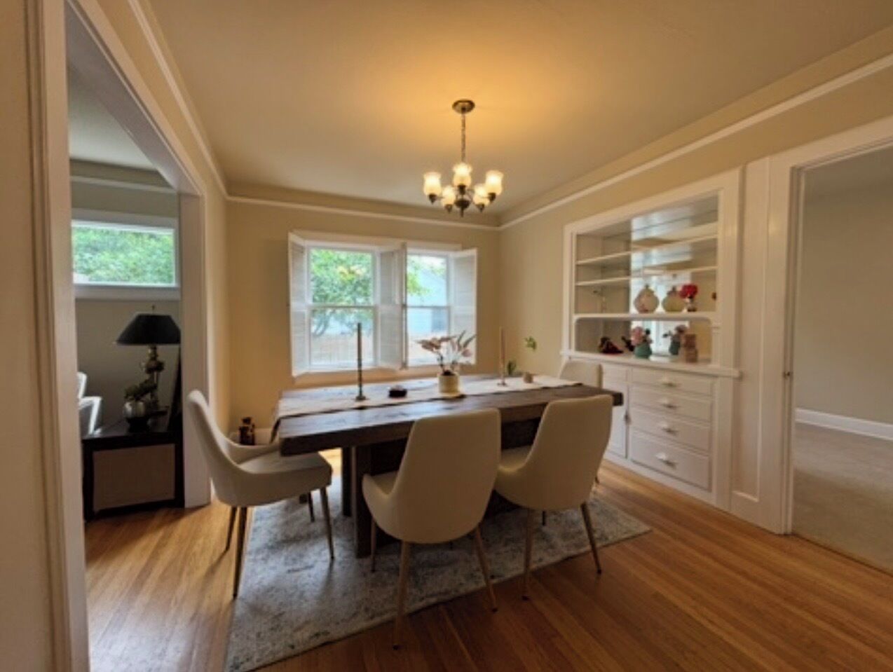 A dining area with a wooden table, four chairs, a chandelier, and a window with a view of greenery.
