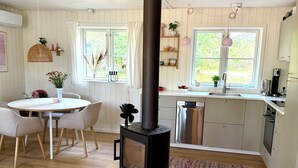 Et moderne køkken med en centralt placeret brændeovn, et spiseområde med et rundt bord og stole, samt udsigt til det fri gennem store vinduer.