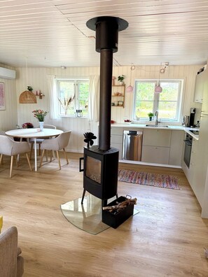 Eine moderne Küche mit einem zentralen Holzofen, einem Essbereich mit rundem Tisch und Stühlen sowie Blick ins Freie durch große Fenster.