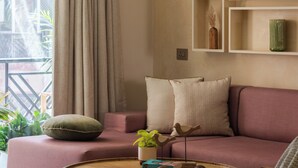 Un soggiorno con un divano, un tavolino rotondo e una poltrona. Sono presenti scaffali con oggetti decorativi e una finestra con tende.