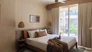 Una camera d'albergo con un letto, una ventola a soffitto, una finestra con tende e un balcone con piante.