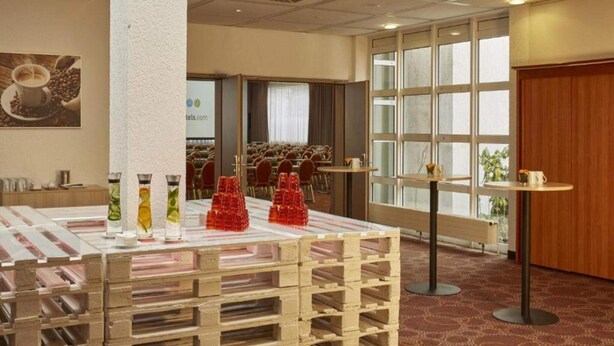 Habitación de hotel con un ventanal grande, una mesa de madera con vasos rojos, una mesita redonda y un pilar.