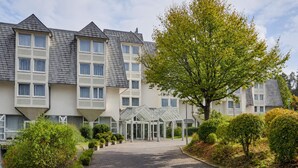 Un hotel de varios pisos con una entrada central, rodeado de jardines y árboles bien cuidados.