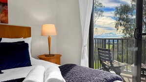 Une chambre à coucher avec un lit, une table de chevet, une lampe, une fenêtre donnant sur un balcon et des arbres.