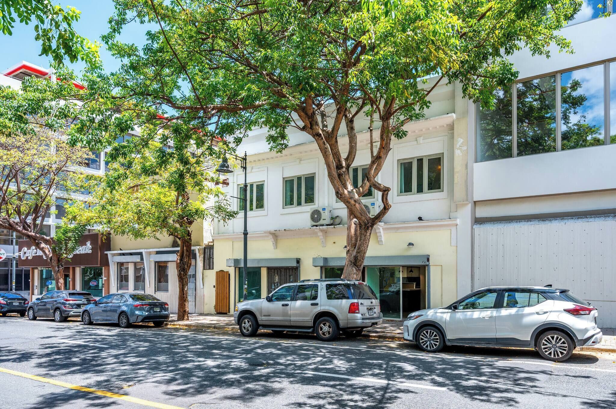 Straßenansicht mit parkenden Autos, einem Baum und einem Gebäude mit weißer Fassade und grünen Fenstern.