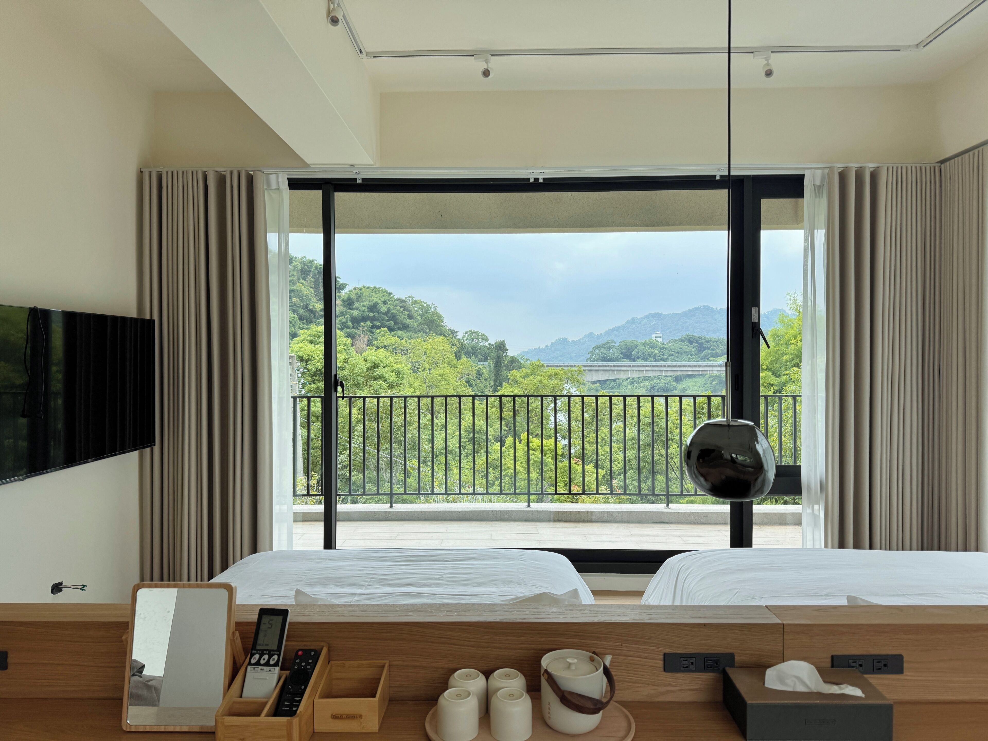 A hotel room with a large window, a bed, a bedside table with a remote control, a mirror, and a view of greenery and a bridge.