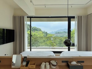 A hotel room with a large window, a bed, a bedside table with a remote control, a mirror, and a view of greenery and a bridge.