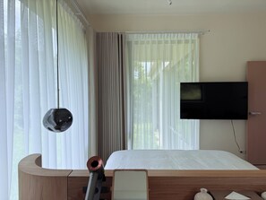Una habitación de hotel con cama, televisor montado y un amplio ventanal con cortinas.