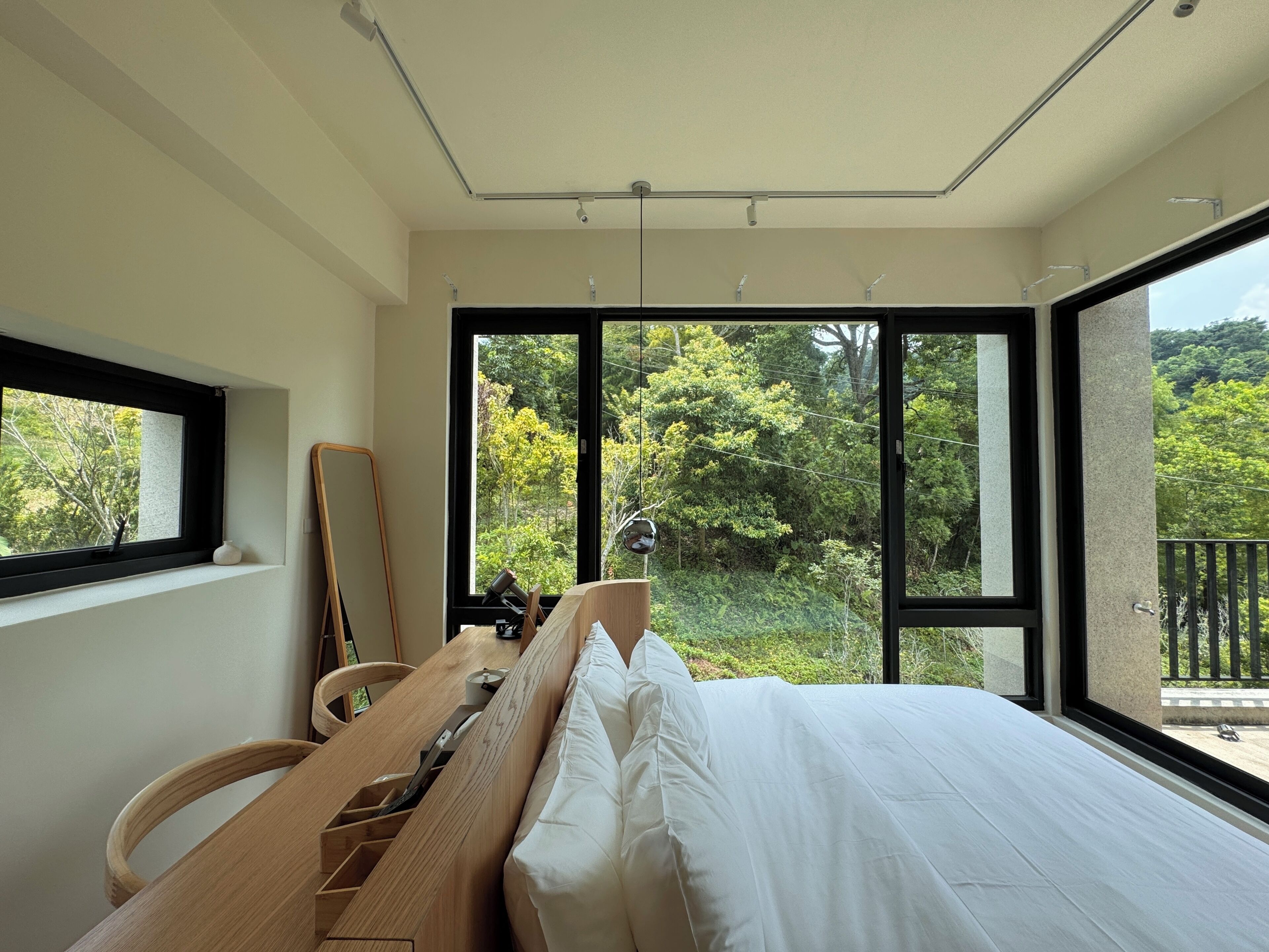 Une chambre avec un grand lit, un bureau en bois et une vue sur la verdure grâce à de grandes fenêtres.