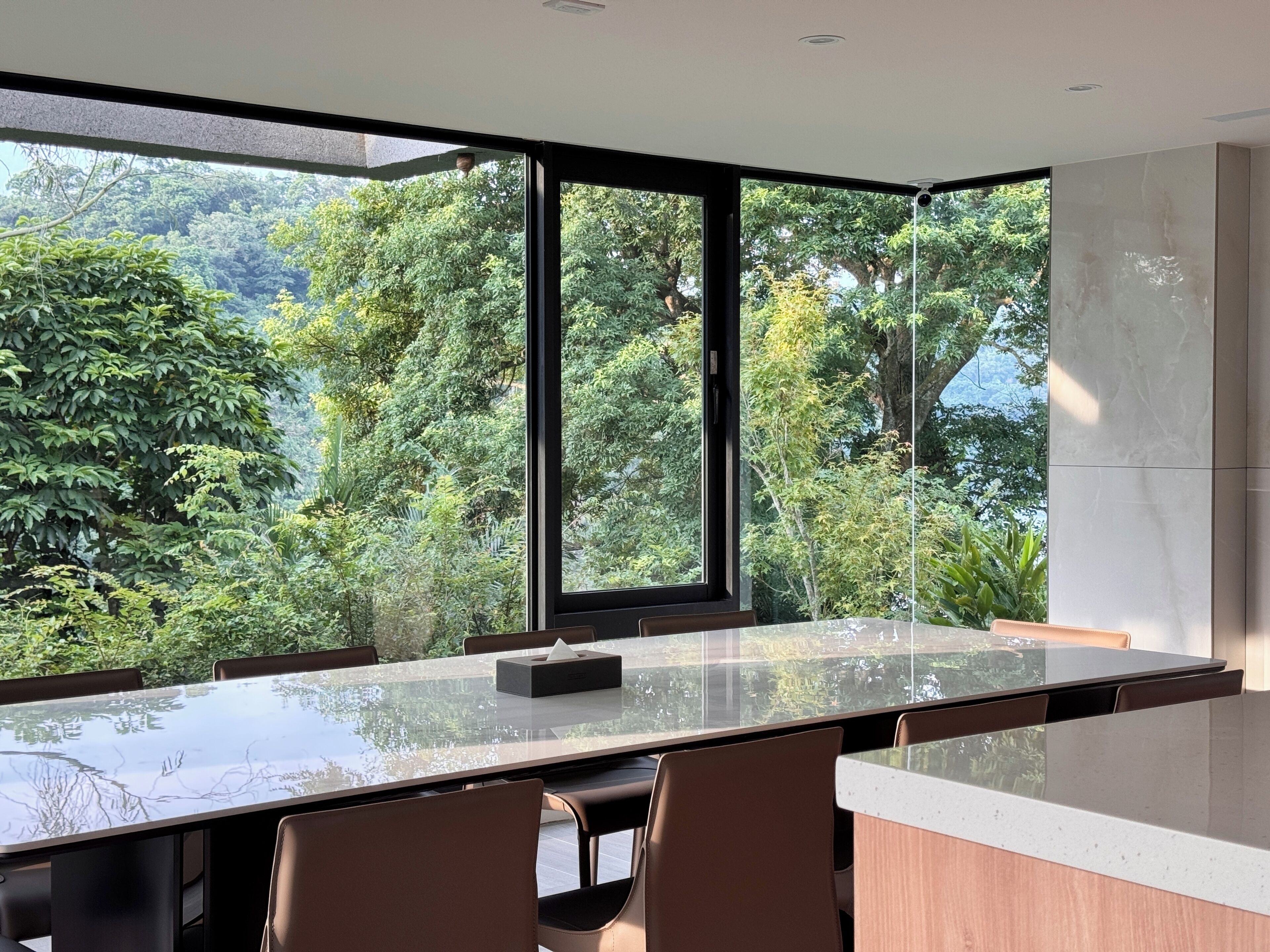 A modern dining area with a glass table and chairs, large windows offering a view of lush greenery.