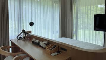 Quarto moderno com escrivaninha de madeira, cadeira, televisão e uma grande janela com cortinas translúcidas.
