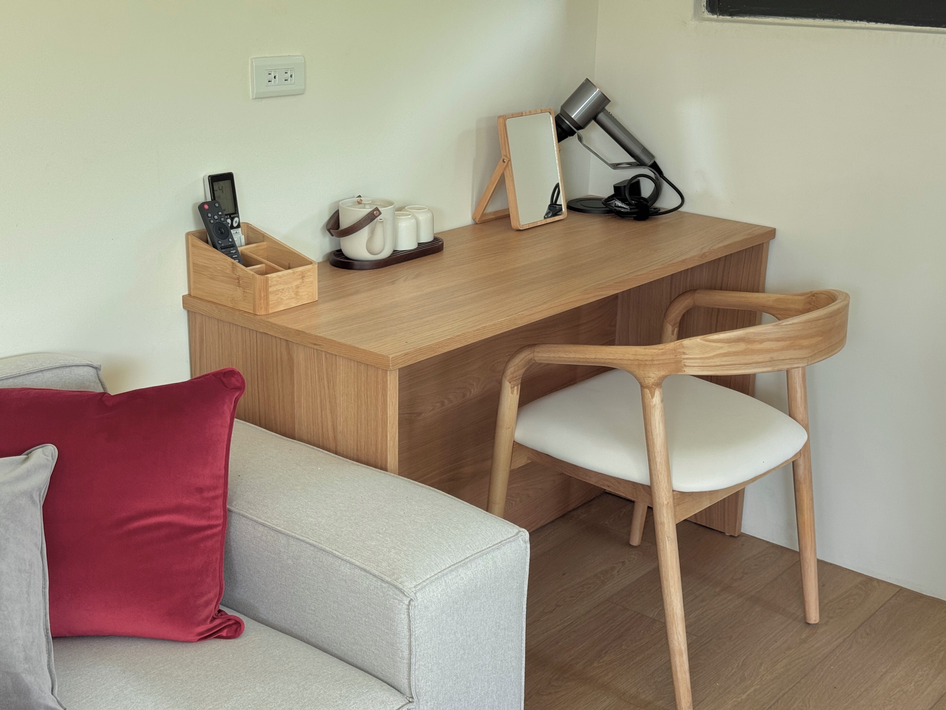 Un espacio habitable compacto con un escritorio de madera, una silla, una mesita de noche con espejo, un secador de pelo y una estación de carga.