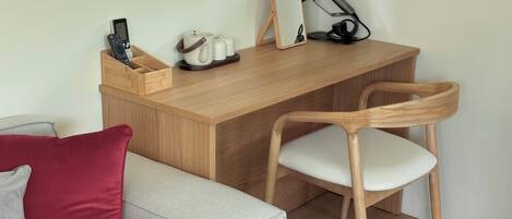 A compact living space with a wooden desk, a chair, a small table with a mirror, a hairdryer, and a charging station.