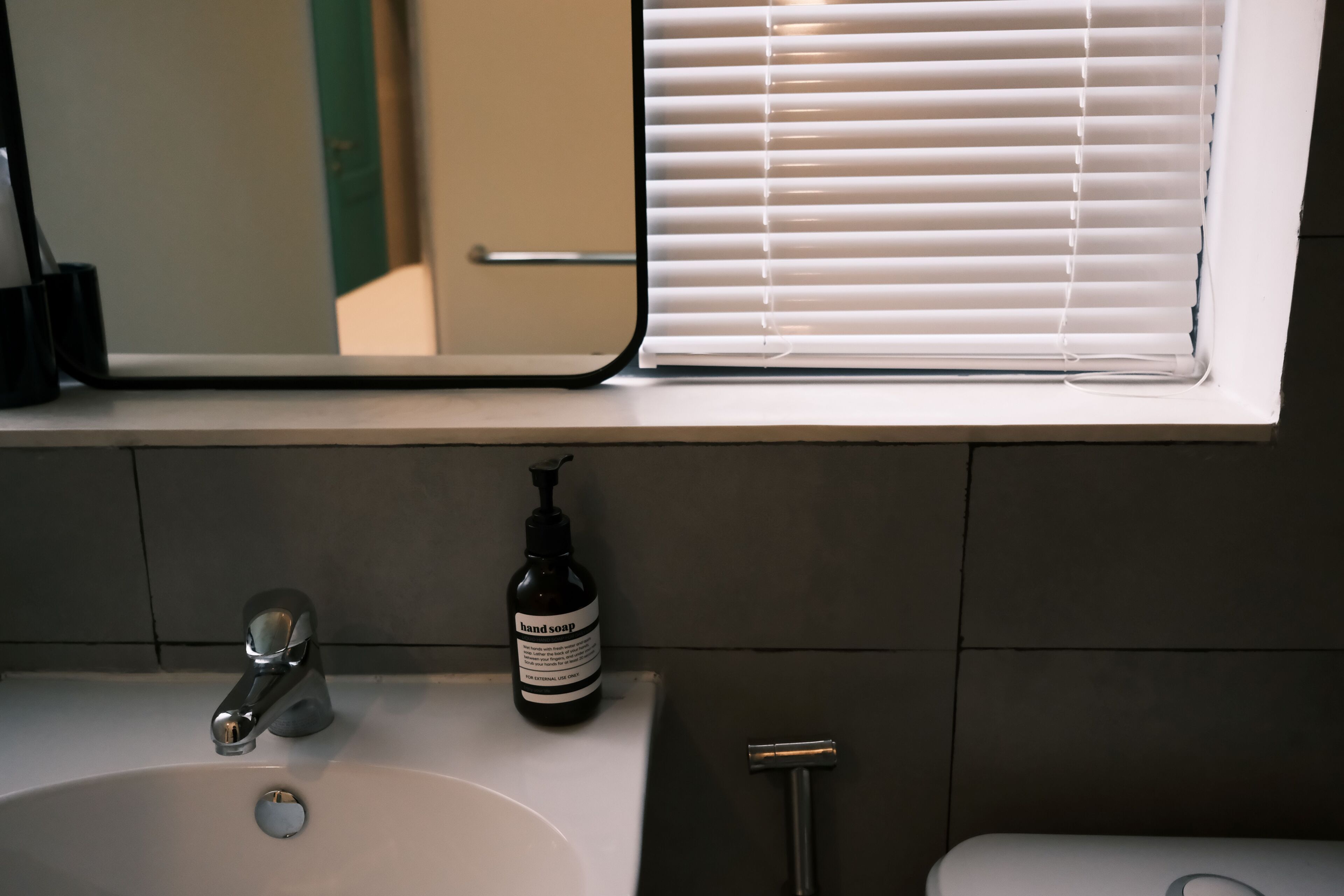 A bathroom with a sink, a hand soap dispenser, a mirror, and a window with blinds.