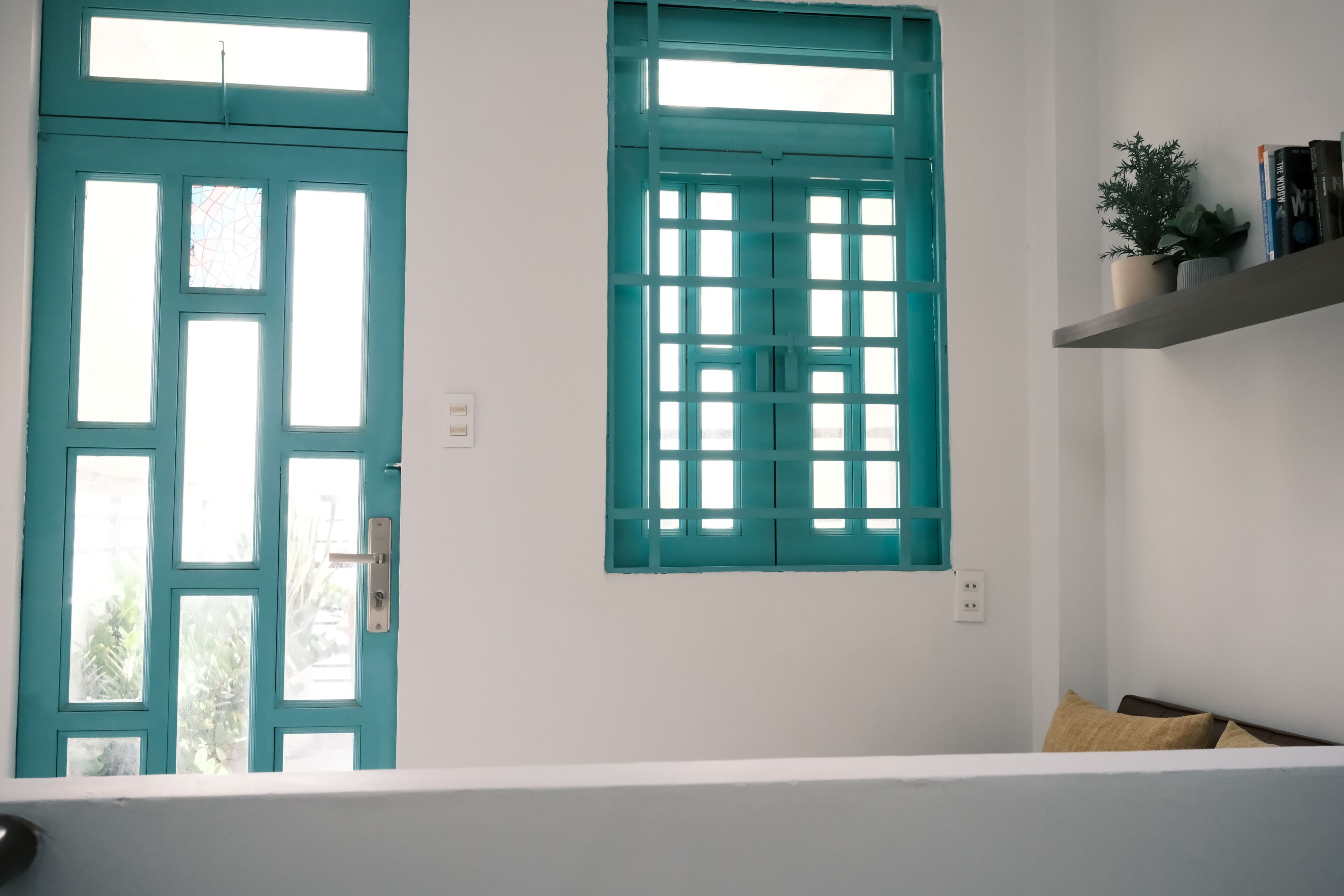 A room with two teal-framed windows, a white wall, a shelf with books and a plant, and a bed with pillows.