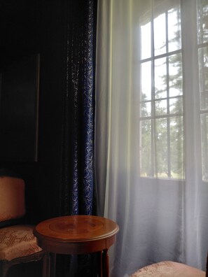 A room with a round wooden table, two patterned chairs, a dark blue curtain with a leaf pattern, and a window with a view of trees.