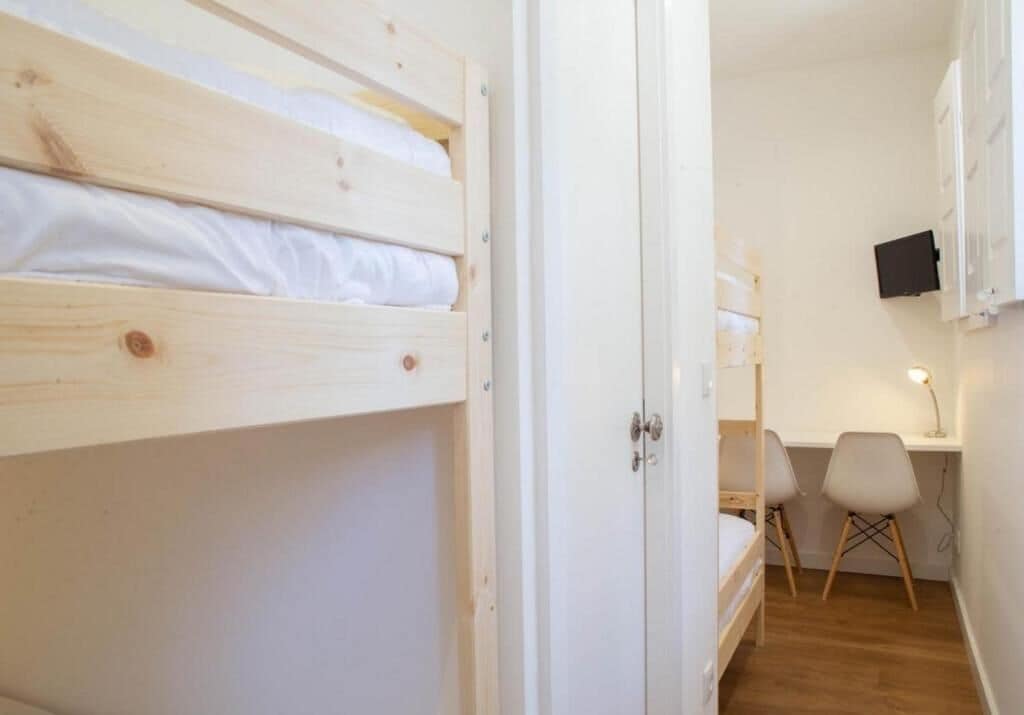 A room with a bunk bed, a desk with a chair, a lamp, and a TV mounted on the wall.