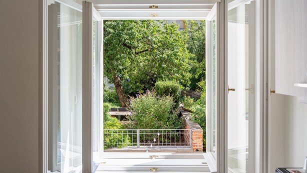 A view through an open window with a white frame, leading to a balcony with a metal railing, surrounded by greenery and a brick wall. - Stylish Design Apartment in Central Prague (Hlavní město Praha)